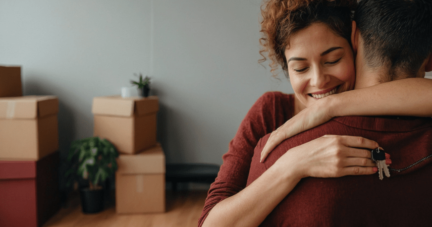Um casal abraçado dentro de seu apartamento novo, enquanto a mulher segura as chaves do imóvel próprio na mão.